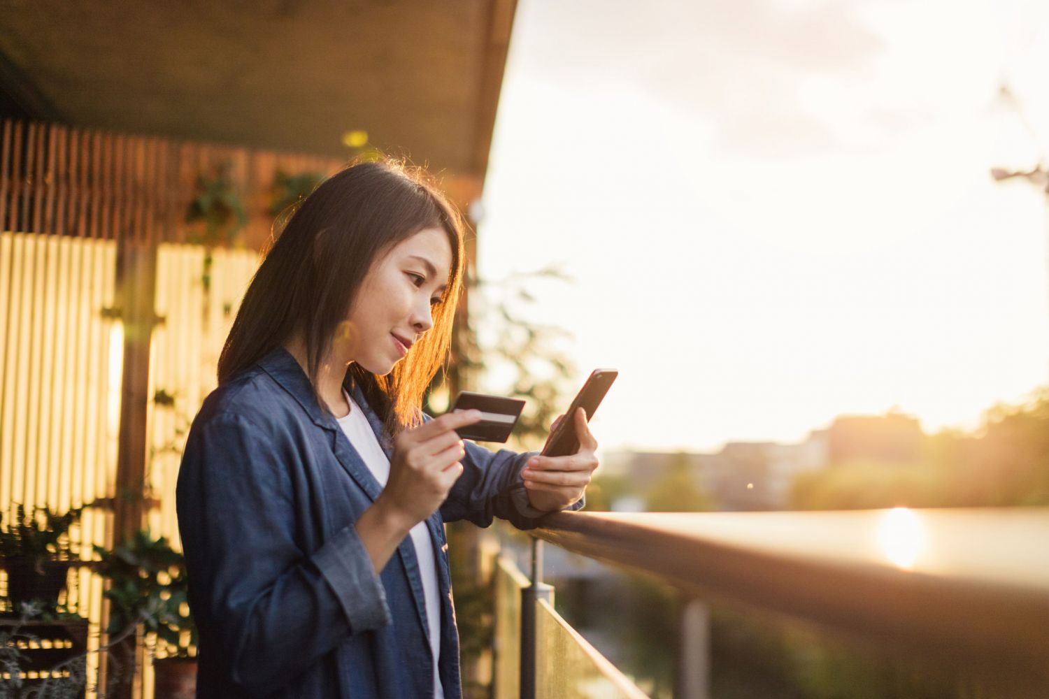 Eine junge Frau steht auf einem Balkon und hält ein Handy sowie eine Kreditkarte in der Hand. 