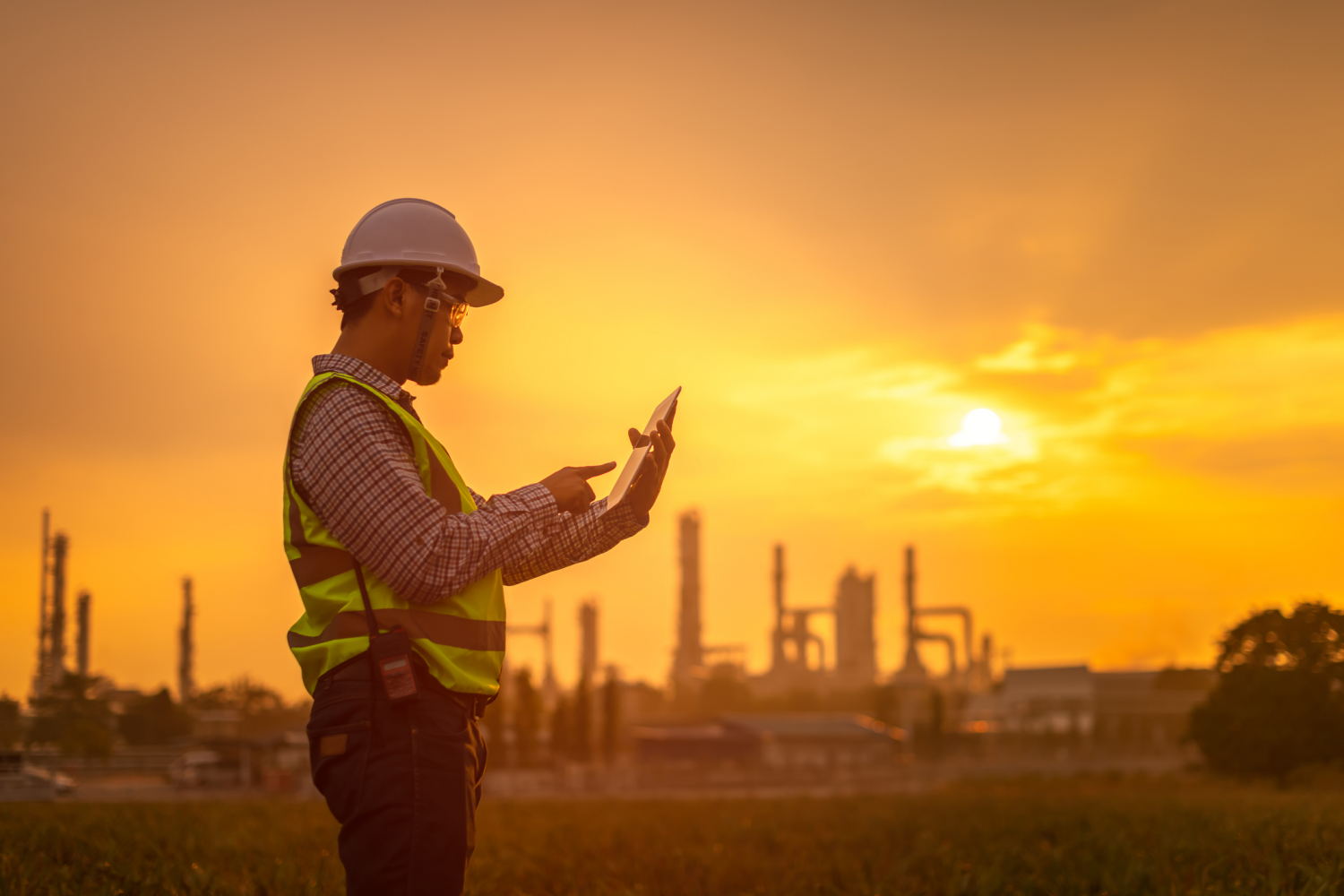 Ein Arbeiter steht bei Sonnenuntergang mit einem Tablet vor einem Industriegelände.